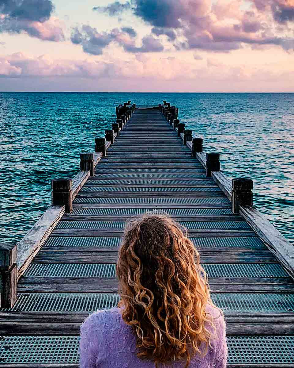 Ein Foto von einer Frau auf einem Steg am wundersch&ouml;nen t&uuml;rkisblauen Meer. Man sieht die r&ouml;tlich-orangenfarbigen Schimmer auf dem Meer beim Sonnenuntergang.