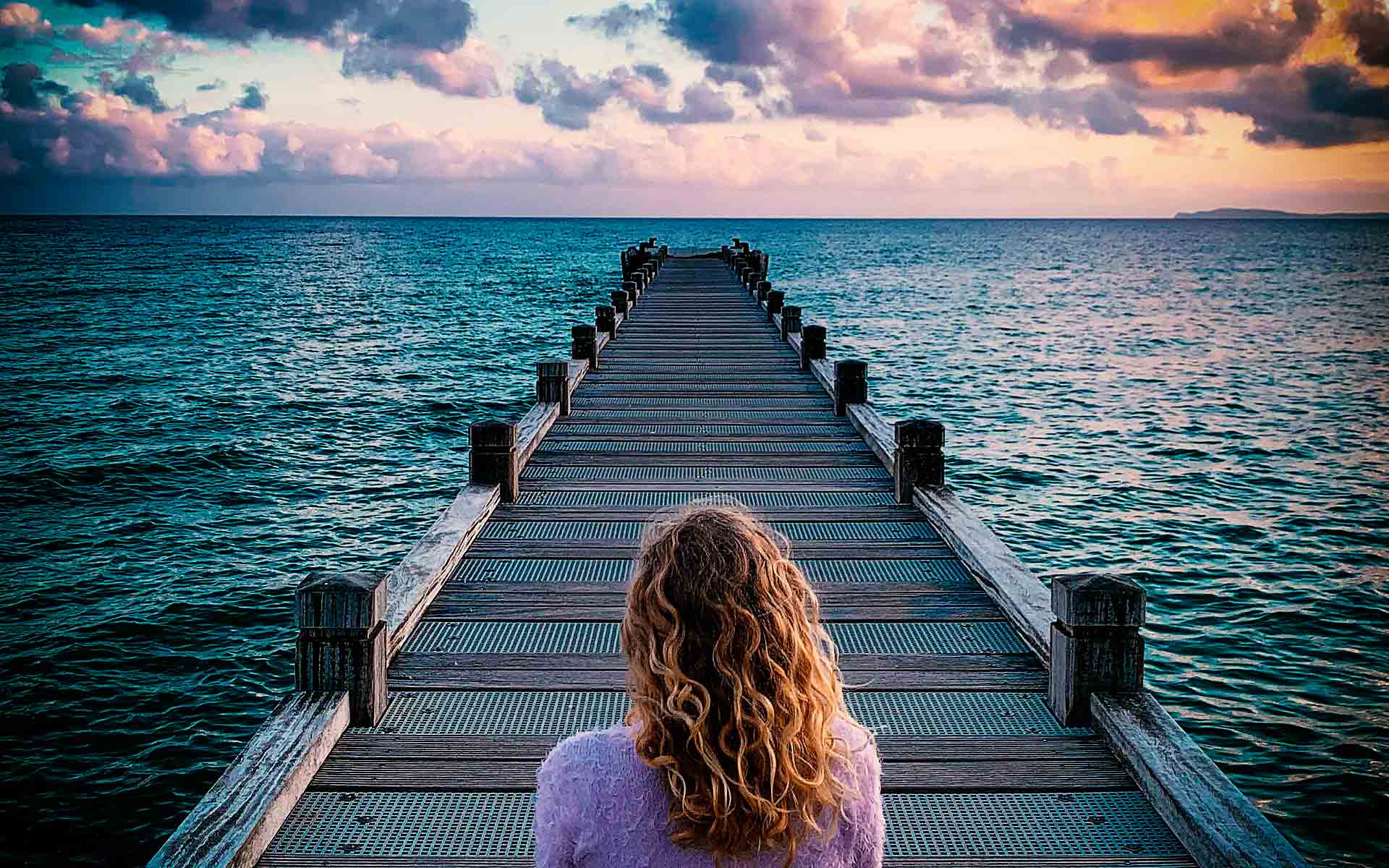Ein Foto von einer Frau auf einem Steg am wunderschönen türkisblauen Meer. Man sieht die rötlich-orangenfarbigen Schimmer auf dem Meer beim Sonnenuntergang.