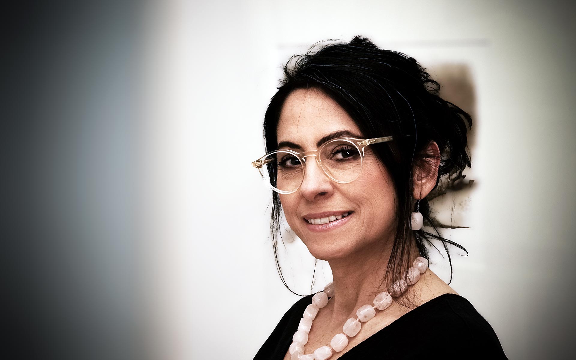 Asli Demirel, Heilpraktikerin für Psychotherapie und Systemische Therapeutin, in ihrer Praxis in Berlin Charlottenburg, stehend, seitlich lächelnd, in schwarzem Kleid, mit Brille, Rosen-Bergkristall-Collier und passenden Ohrringen.“