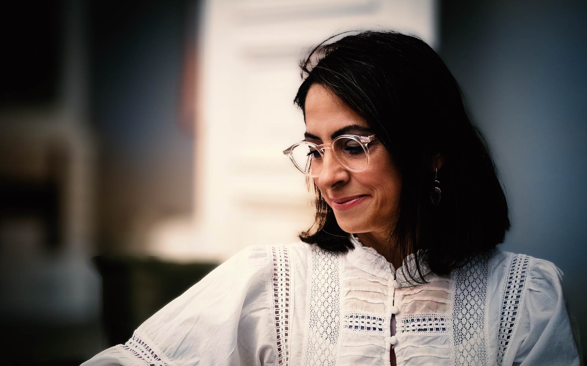 Asli Demirel, Systemische Therapeutin und Heilpraktikerin für Psychotherapie, lächelnd, sitzend an ihrem Schreibtisch in Berlin Mariendorf, in weißem Kleid und Brille.