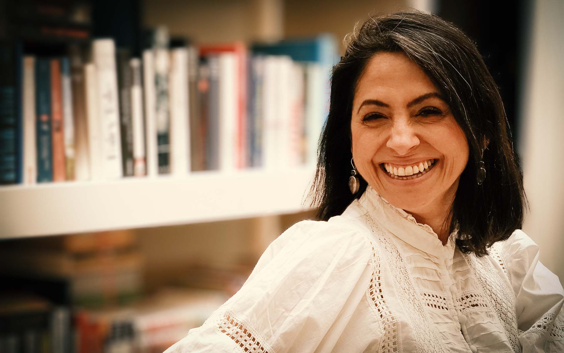 Ein Foto von Asli Demirel, Systemische Therapeutin in Berlin, lächelnd, sitzend in einem weißen Kleid, mit einem weißen Bücherregal im Hintergrund mit vielen Büchern.