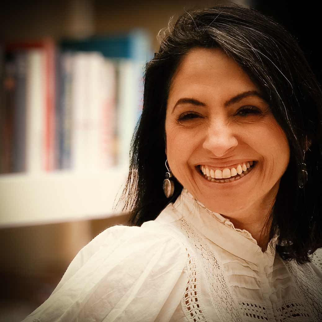 Ein Foto von Asli Demirel, Systemische Therapeutin in Berlin, lächelnd, sitzend in einem weißen Kleid, mit einem weißen Bücherregal im Hintergrund mit vielen Büchern.