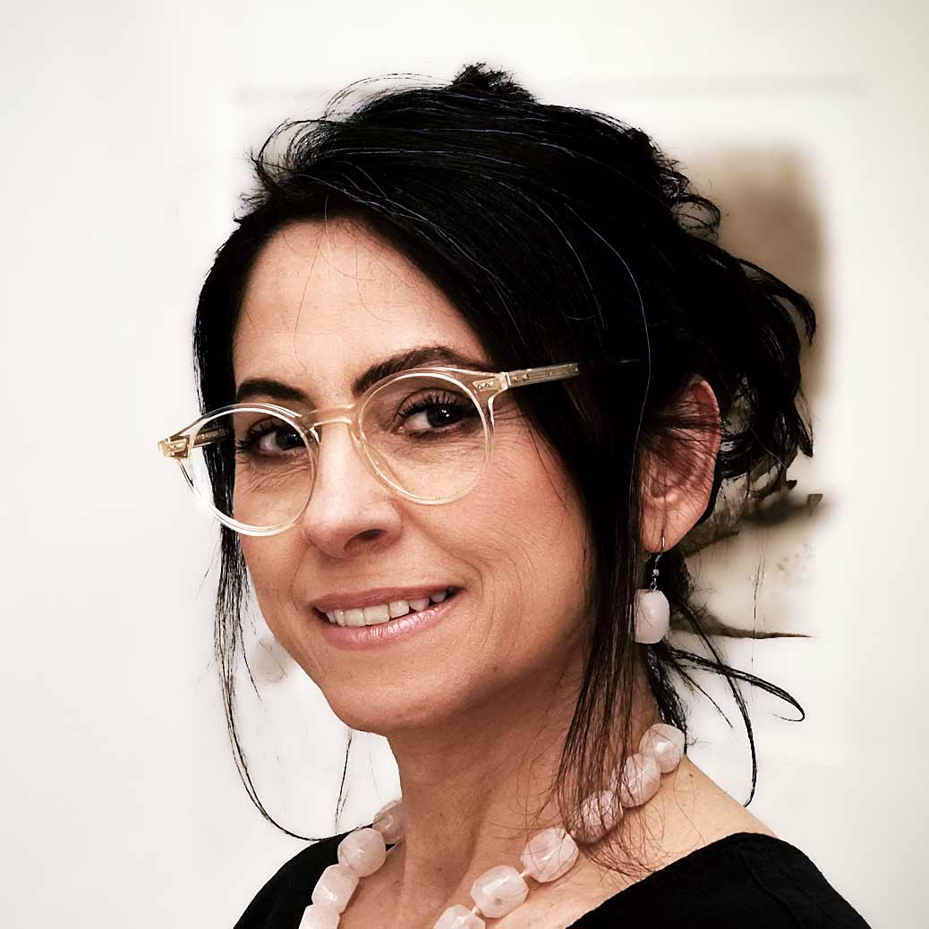 Asli Demirel, Systemische Therapeutin, in ihrer Praxis in Berlin Charlottenburg, lächelnd, in schwarzem Kleid, mit Brille, Rosen-Bergkristall-Collier und passenden Ohrringen.